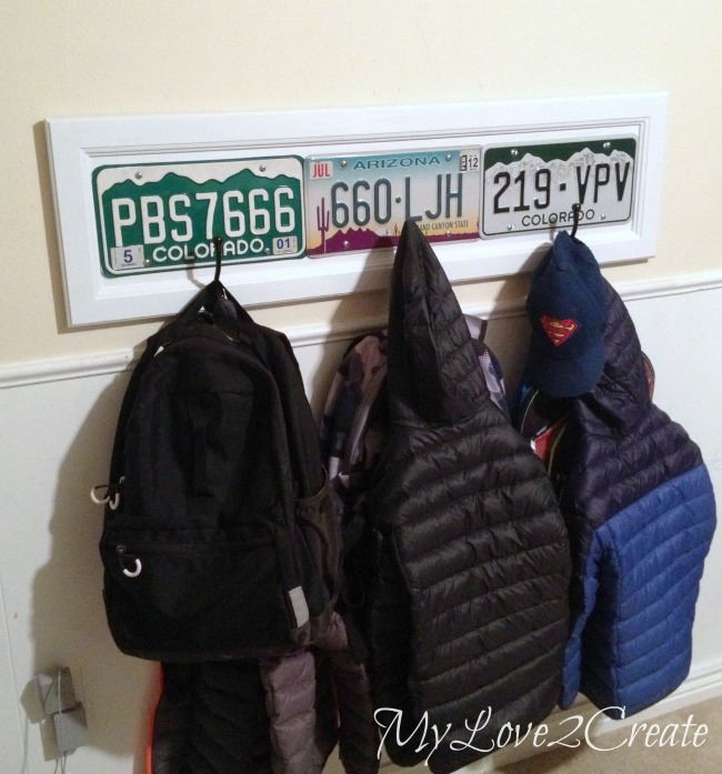 Wall-mounted coat and backpack rack made from a repurposed cupboard door, decorated with vintage license plates from Colorado and Arizona, and featuring hooks for hanging jackets, backpacks, and hats.