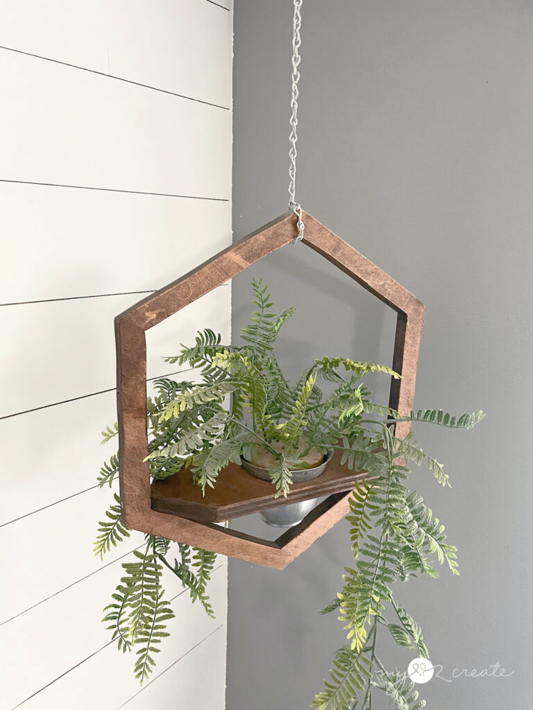 Hexagon-shaped wooden hanging planter with cascading greenery, suspended by a chain against a white shiplap wall.