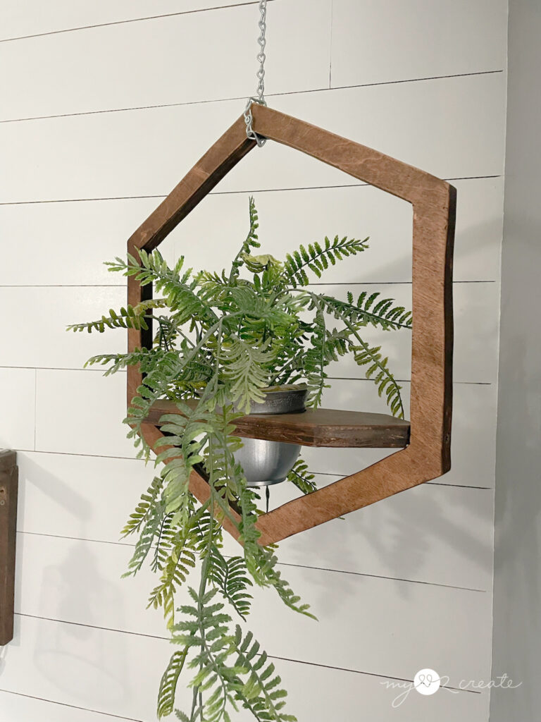 Hexagon-shaped wooden hanging planter with cascading greenery, suspended by a chain against a white shiplap wall.