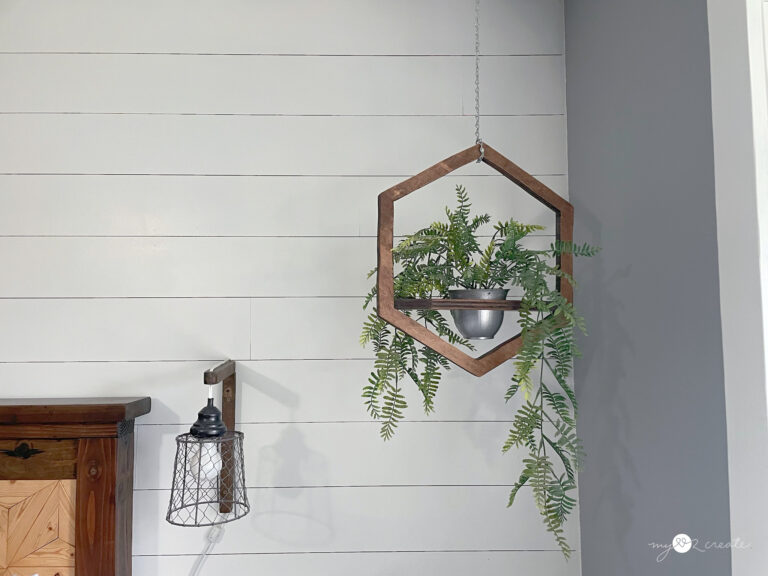 Hexagon-shaped wooden hanging planter with cascading greenery, suspended by a chain against a white shiplap wall.