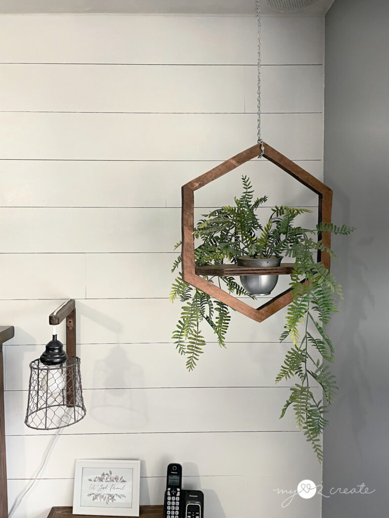 Hexagon-shaped wooden hanging planter with cascading greenery, suspended by a chain against a white shiplap wall.