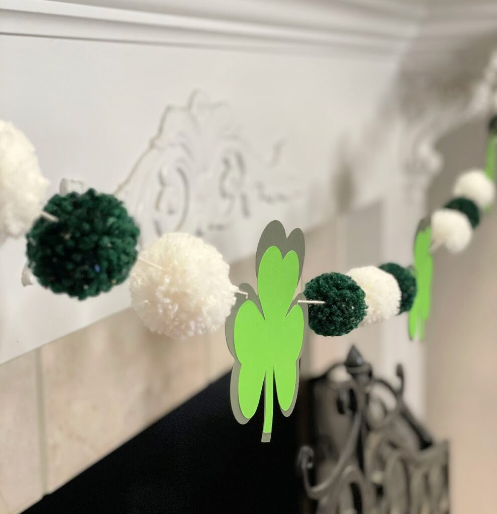 A close-up of a "Get Lucky" St. Patrick's Day garland featuring green shamrocks and alternating dark green and white pom-poms, strung across a white fireplace mantel.