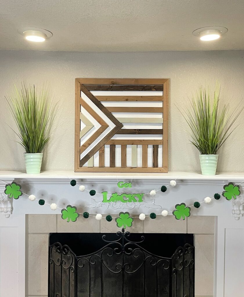 A festive St. Patrick's Day mantel decoration featuring a "Get Lucky" shamrock garland with green and white pom-poms, hung above a black wrought iron fireplace. The mantel is adorned with two green potted plants and a geometric wooden wall art piece.