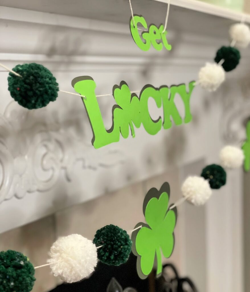 A close-up of a "Get Lucky" St. Patrick's Day garland featuring green shamrocks and alternating dark green and white pom-poms, strung across a white fireplace mantel.