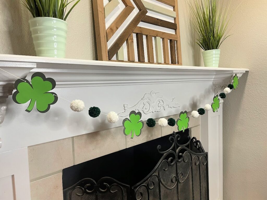 A festive St. Patrick's Day mantel decoration featuring a "Get Lucky" shamrock garland with green and white pom-poms, hung above a black wrought iron fireplace. The mantel is adorned with two green potted plants and a geometric wooden wall art piece.