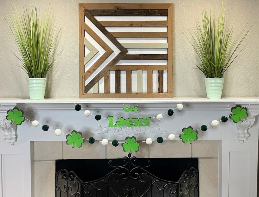 A festive St. Patrick's Day mantel decoration featuring a "Get Lucky" shamrock garland with green and white pom-poms, hung above a black wrought iron fireplace. The mantel is adorned with two green potted plants and a geometric wooden wall art piece.