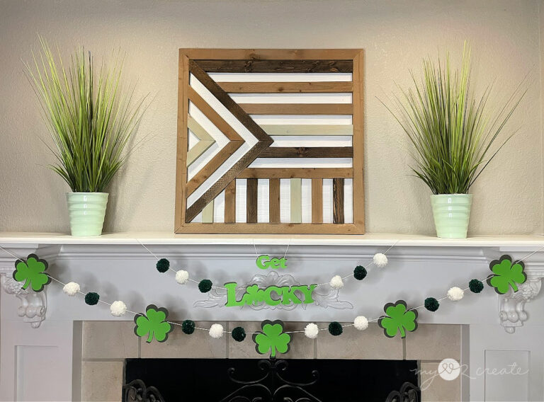 A St. Patrick's Day mantel decoration featuring a shamrock garland with green and white pom-poms, a geometric wooden wall art piece, and two green potted plants.