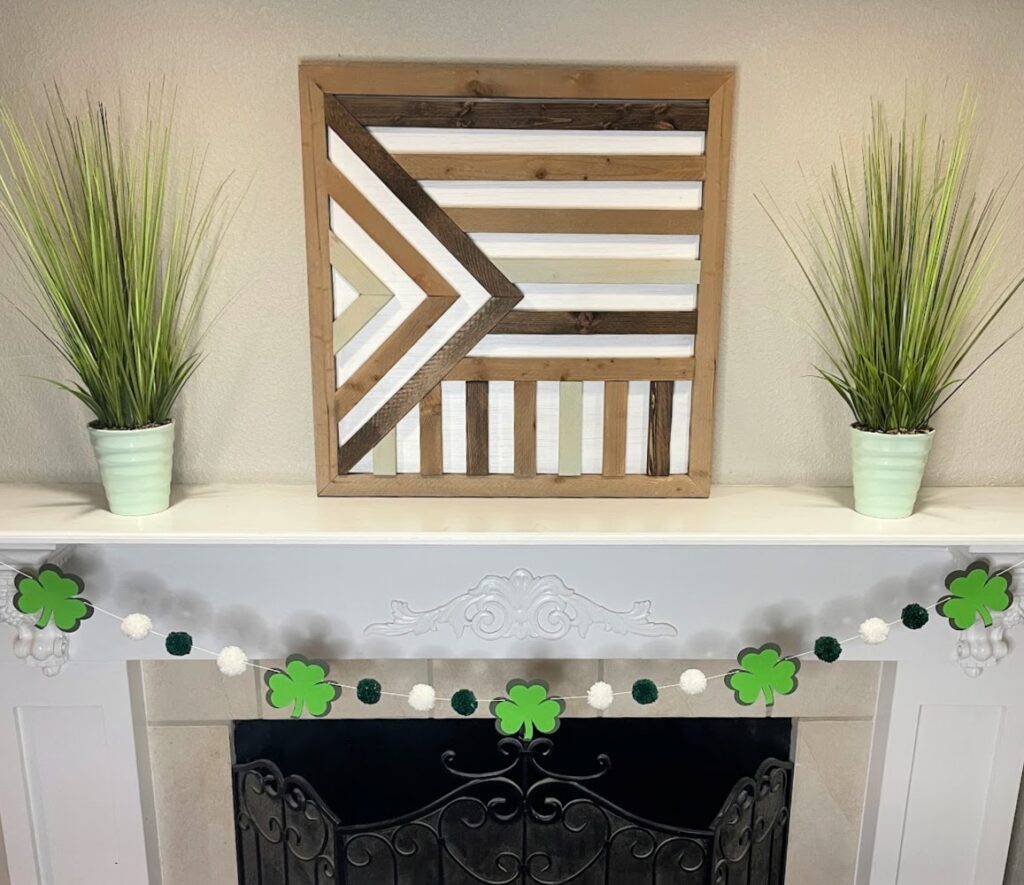 A St. Patrick's Day mantel decoration featuring a shamrock garland with green and white pom-poms, a geometric wooden wall art piece, and two green potted plants.