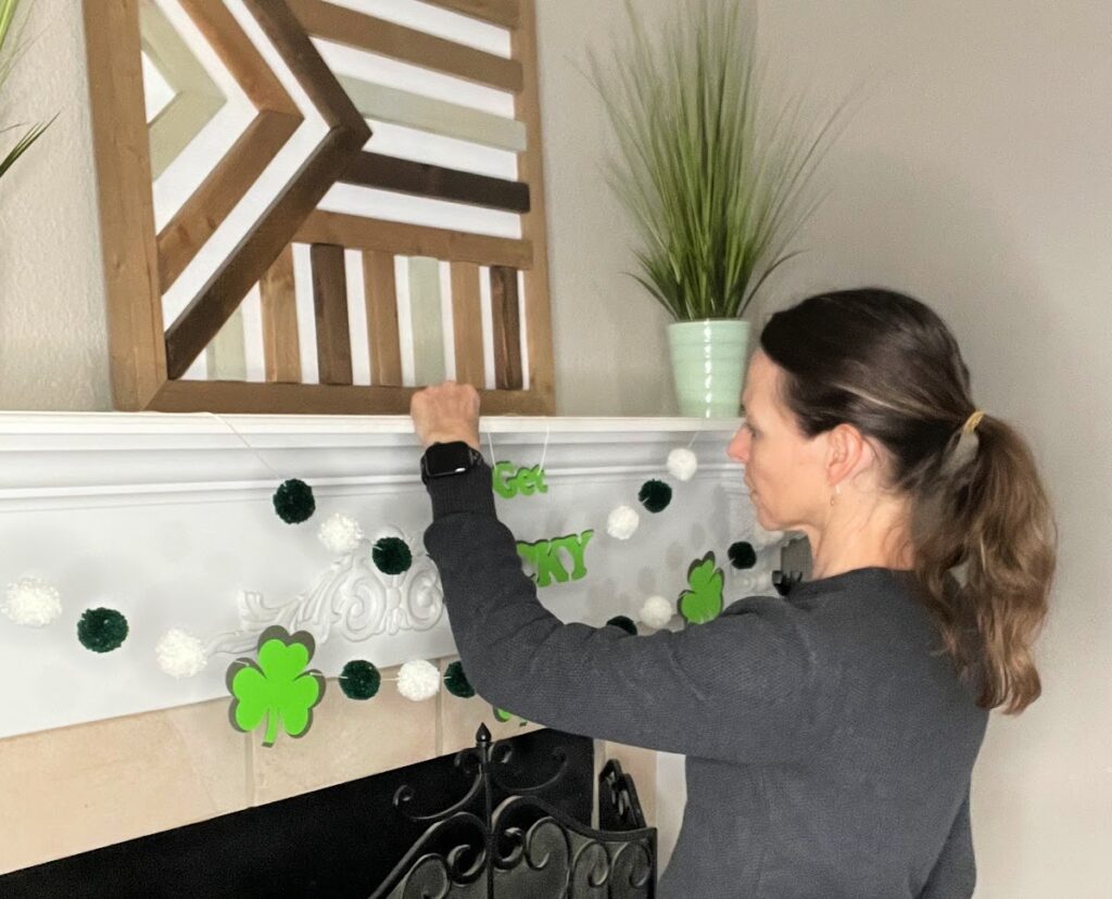 A woman in a dark sweater decorates a white fireplace mantel by hanging a St. Patrick's Day garland with green shamrocks, pom-poms, and the words "Get Lucky."