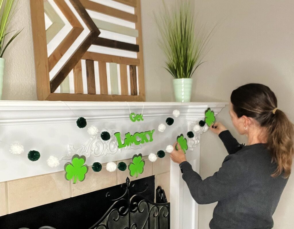 A woman adjusts a festive "Get Lucky" shamrock garland on a white fireplace mantel, ensuring even spacing of decorations.