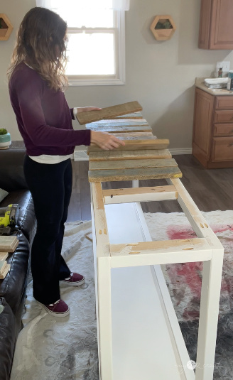 Woman arranging reclaimed wood planks on a white table frame in a bright indoor workspace, preparing for a DIY furniture project.