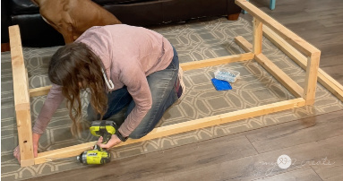 A woman kneeling on a patterned rug while assembling a large wooden frame. She is using a cordless drill to secure the structure. A bag of screws is nearby, with a couch and wooden flooring in the background.