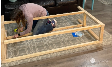 A woman kneeling on a patterned rug while assembling a large wooden frame. She is using a cordless drill to secure the structure. A bag of screws is nearby, with a couch and wooden flooring in the background.