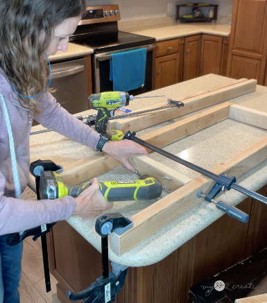 A woman using a Ryobi cordless tool to sand or smooth wooden boards clamped to a kitchen island. A drill and other tools are on the counter. The kitchen has wooden cabinets, stainless steel appliances, and a beige countertop.