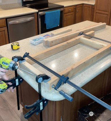 A woodworking project in progress on a kitchen island, with wooden boards clamped together using bar clamps. A Ryobi cordless drill is being used to drive screws into the frame. The workspace includes a measuring tape, screws, and extra wood pieces, with a kitchen setting in the background.