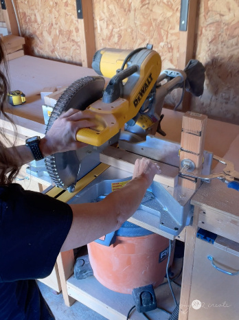 Person using a DeWalt miter saw in a woodworking workshop, with a stop block clamped for precision cutting. A shop vacuum is positioned under the saw for dust collection.