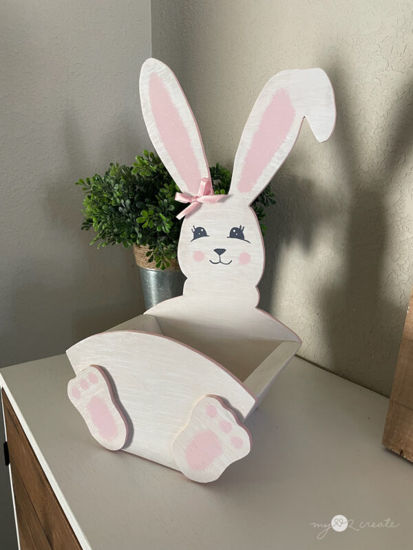A wooden Easter Bunny crate, painted white with pink details, placed on a white tabletop with a green potted plant in the background.