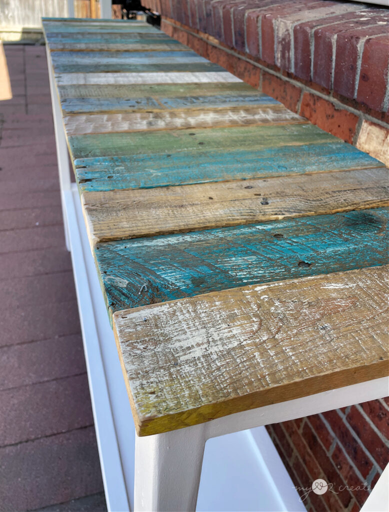 A close-up of a console table's rustic reclaimed wood top, featuring distressed planks in shades of blue, green, and natural wood tones.