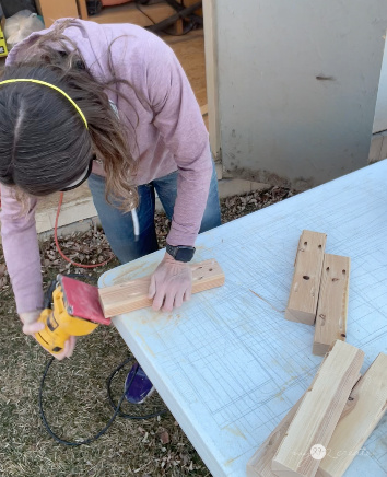 Woman using a yellow power sander to smooth a wooden block on an outdoor table, with pre-cut wood pieces and a shed in the background.