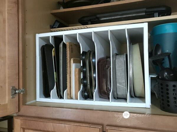 A white wooden vertical storage organizer inside a kitchen cabinet, neatly holding various baking sheets, cutting boards, muffin tins, and other bakeware.