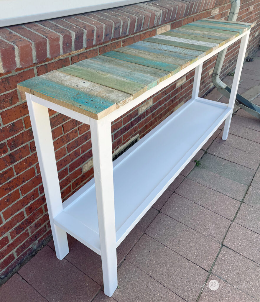 White-framed console table with a reclaimed wood slat top in blue, green, and natural hues, set against a red brick wall on a paved patio.