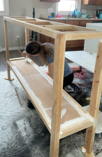 Woman kneeling while painting the lower shelf of a wooden table frame in an indoor workspace with a kitchen in the background.