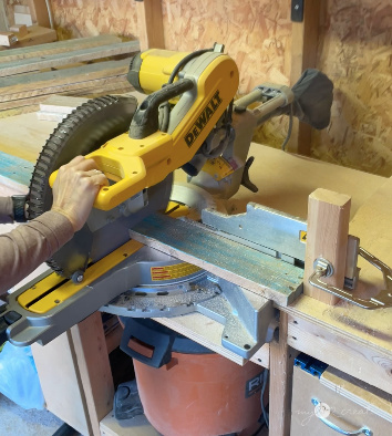 Close-up of a person using a DeWalt miter saw to cut a piece of reclaimed wood in a woodworking workshop with tools and a shop vacuum visible.