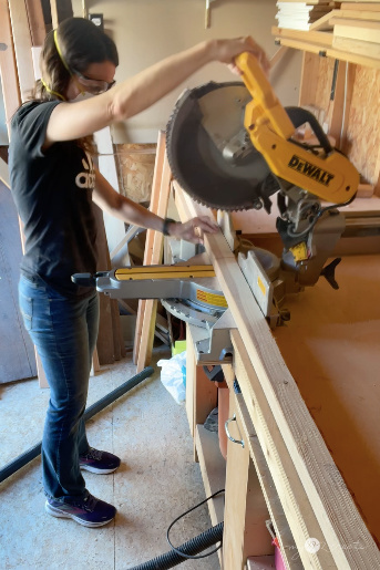 Person using a DeWalt miter saw in a woodworking workshop, with a stop block clamped for precision cutting. A shop vacuum is positioned under the saw for dust collection.