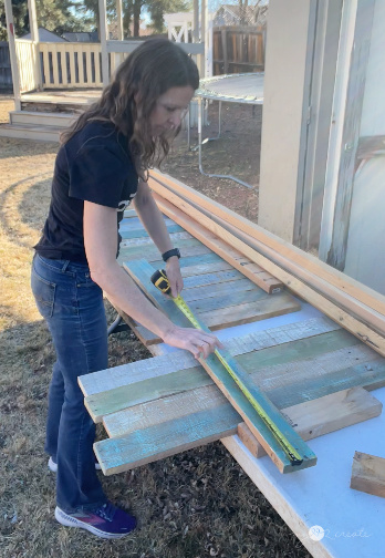 A woman measuring and marking reclaimed wood planks on a table outdoors using a tape measure and a straight edge. The wood has a rustic, weathered look with faded blue, green, and white paint. A wooden porch and a trampoline are in the background.