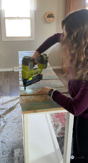 Woman using a Ryobi nail gun to attach reclaimed wood planks to a white table frame in a well-lit indoor workspace.