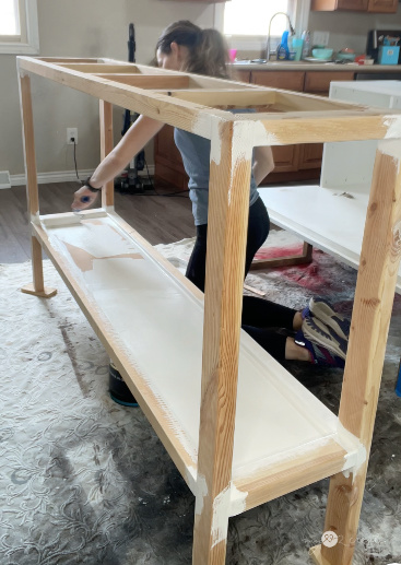 Woman kneeling while painting a wooden table frame white in an indoor workspace, with a kitchen in the background.