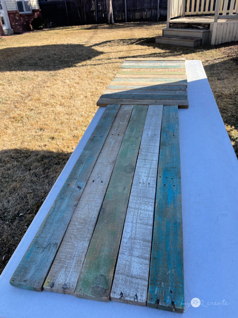 Reclaimed wood planks with faded blue, green, and white paint laid out on a table outdoors, prepared for a DIY woodworking project. A wooden porch and a yard with dried grass are in the background.