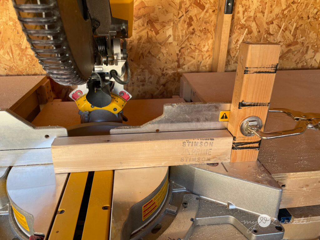 Close-up of a miter saw cutting a 2x4 board, with a stop block clamped in place for consistent cuts. The saw is set up in a woodworking workshop with sawdust and tools in the background.