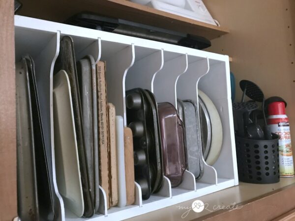 A white wooden vertical storage organizer inside a kitchen cabinet, neatly holding various baking sheets, cutting boards, muffin tins, and other bakeware.