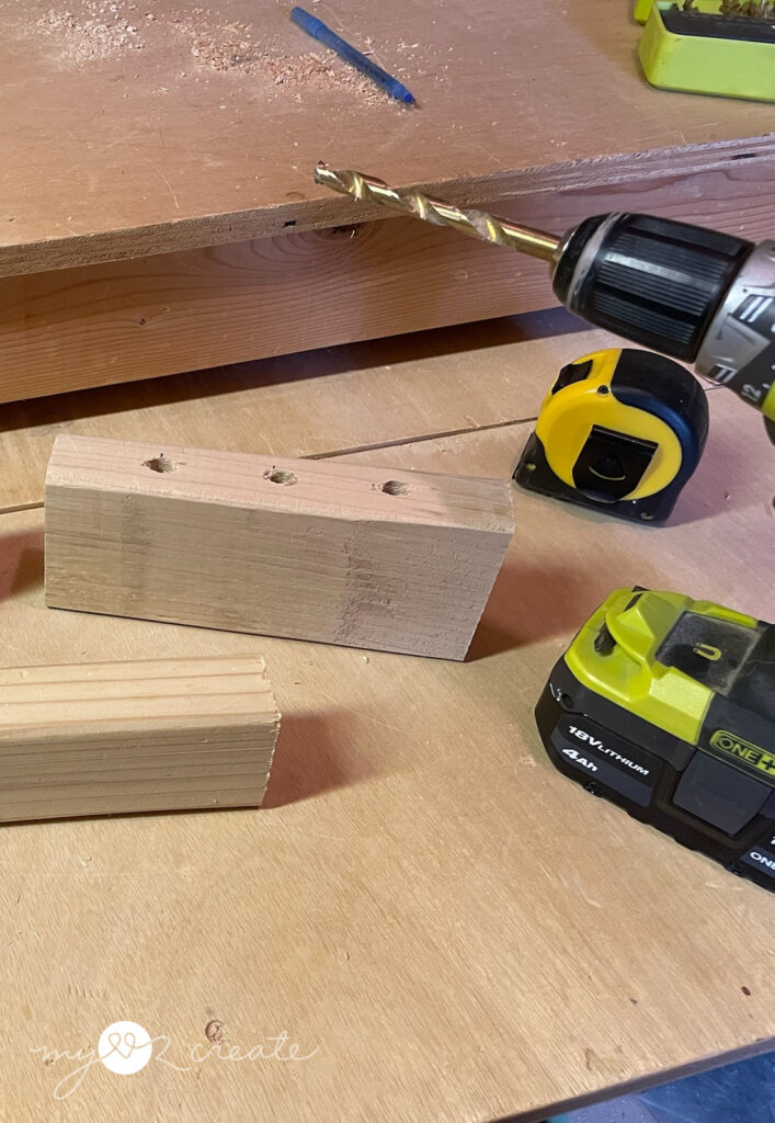 A close-up of a woodworking project in progress, showing a power drill, a tape measure, and a piece of wood with three drilled holes on a workbench.