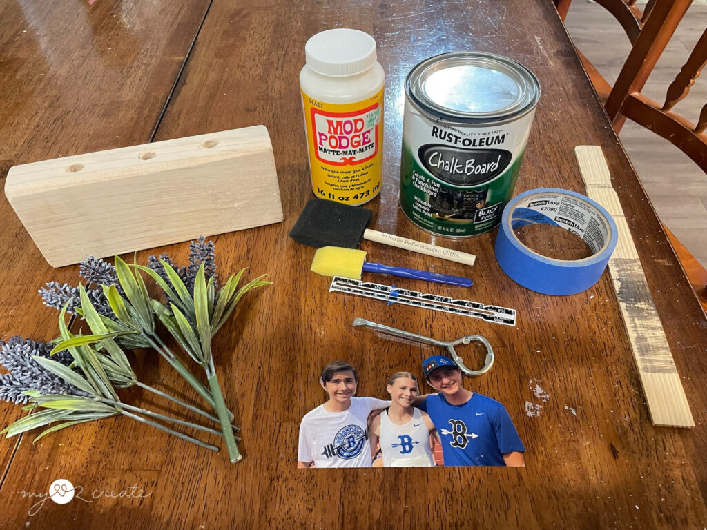 Craft project supplies laid out on a wooden table, including a drilled wood block, faux lavender stems, Mod Podge, chalkboard paint, painter's tape, foam brush, photo cutout, and other crafting tools.