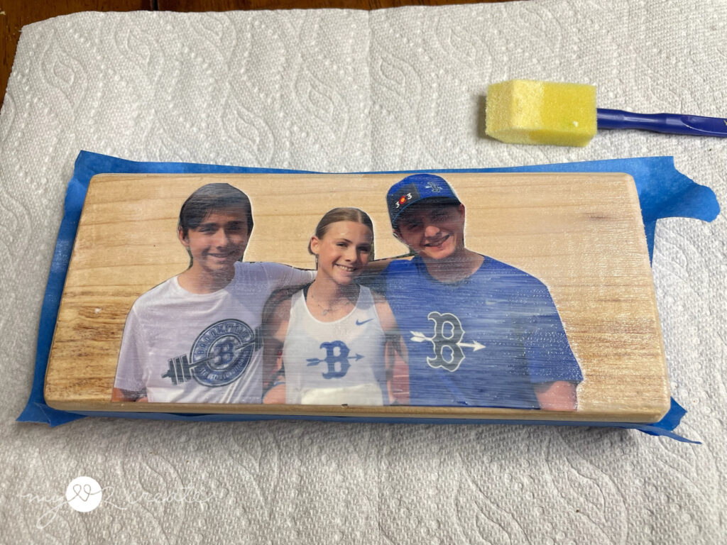 A printed photo of three smiling teenagers adhered to a wooden block using Mod Podge, with painter’s tape around the edges and a foam brush nearby.